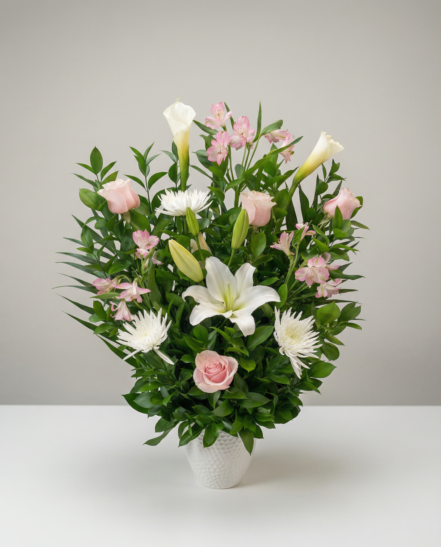 Arrangement délicat composé de lys blancs, de roses pastel, d’alstroemérias rosées et de fleurs blanches texturées. Une création florale tendre et lumineuse, idéale pour offrir réconfort, douceur et compassion lors d’un moment de recueillement.