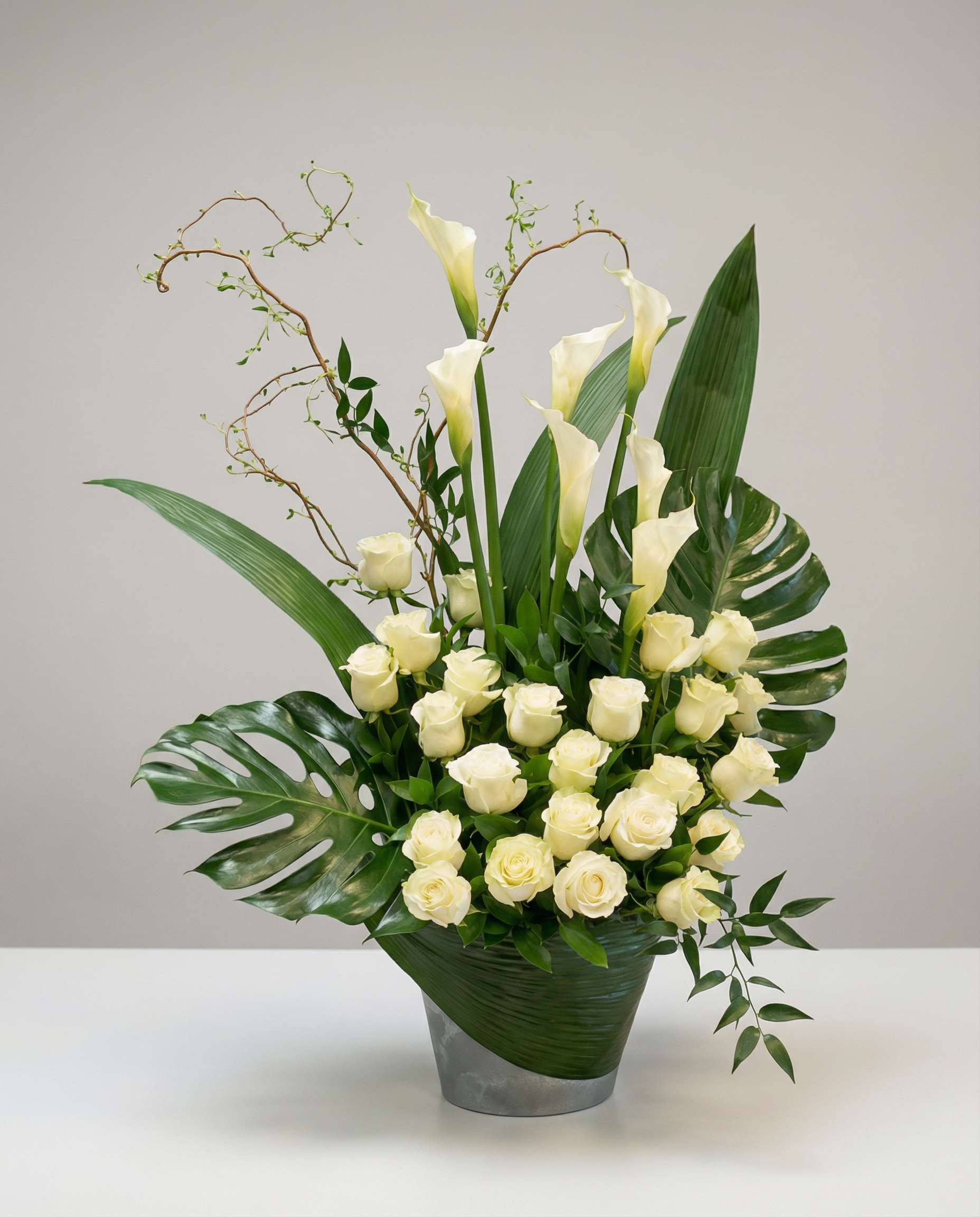 Arrangement raffiné composé de roses blanches et de callas élancés, sublimé par un feuillage tropical et des branches décoratives. Présentée dans un contenant gris moderne, cette corbeille funéraire offre un hommage sobre, élégant et apaisant, idéal pour exprimer respect et recueillement.